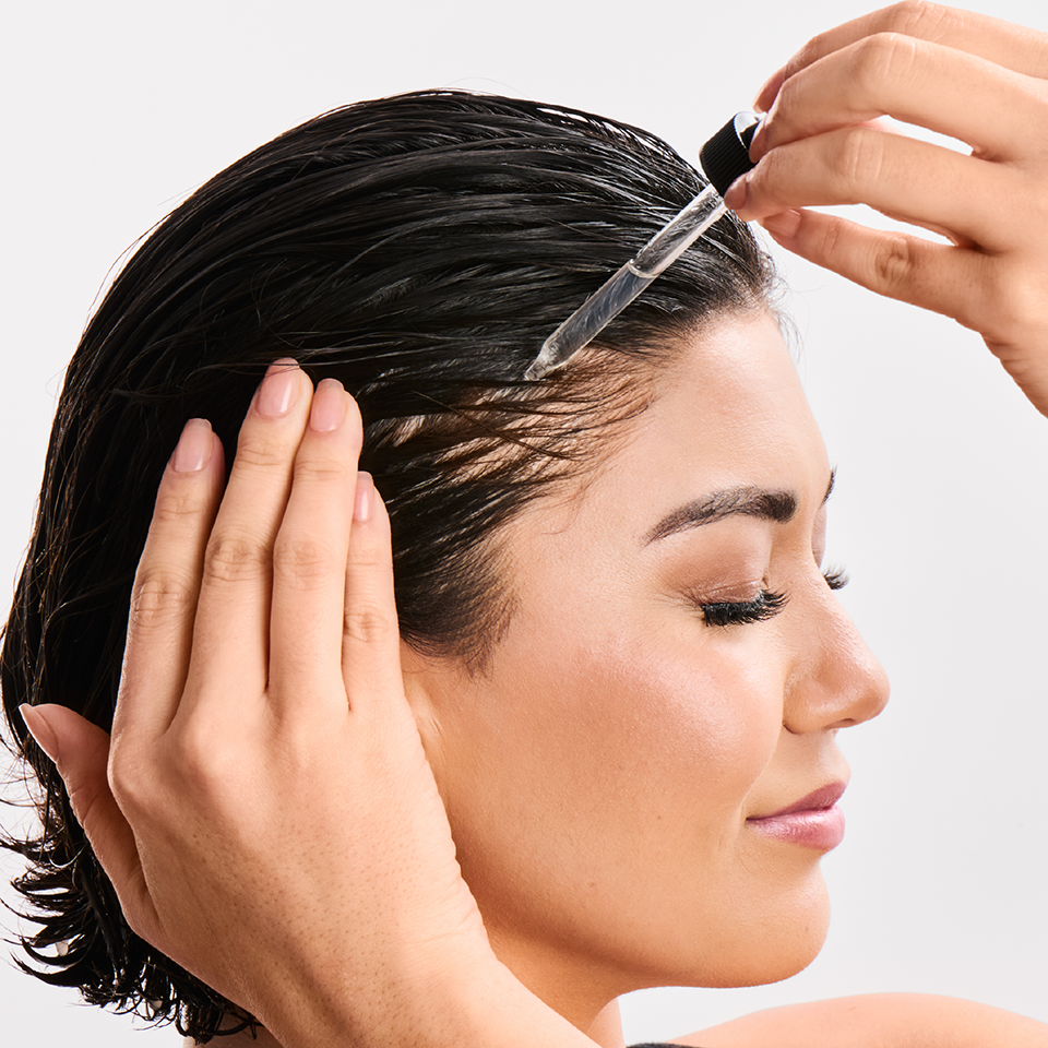 Woman applying hair product to her hair with a brush against a neutral background